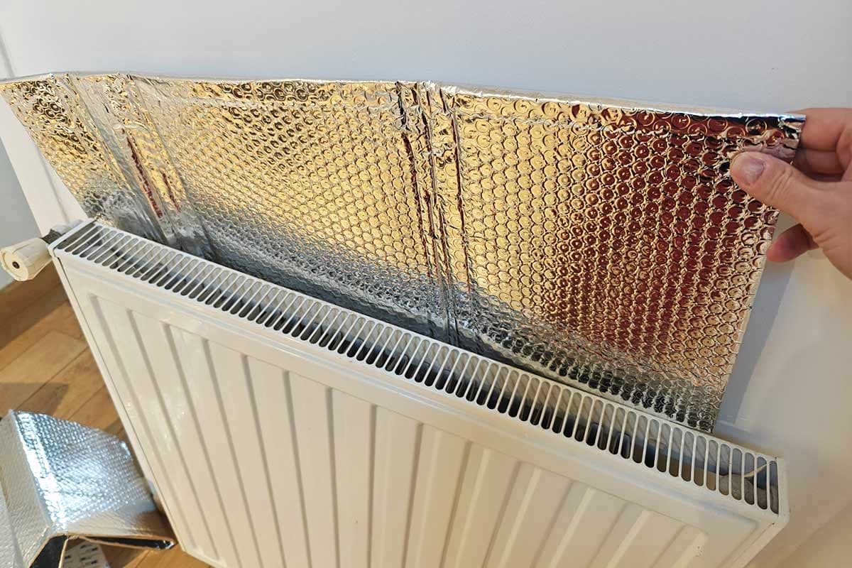 Main glissant un panneau isolant réfléchissant en aluminium derrière un radiateur à eau blanc fixé sur un mur — isolation derrière radiateur
