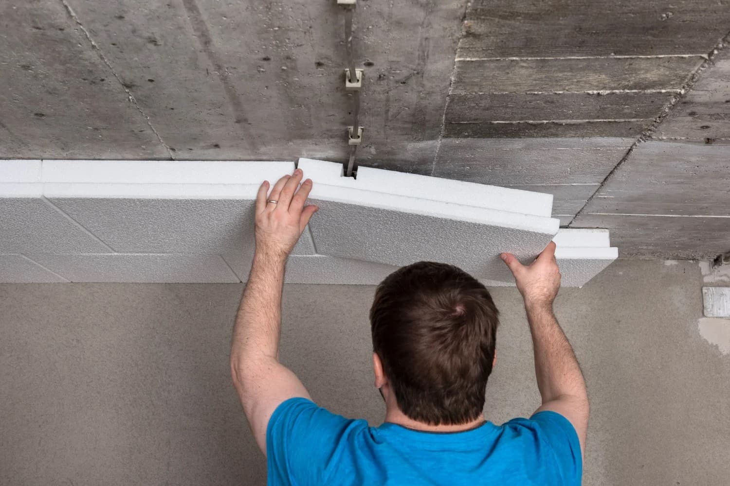 Homme en t-shirt bleu posant des panneaux de polystyrène expansé blanc bord à bord sous un plafond en béton brut