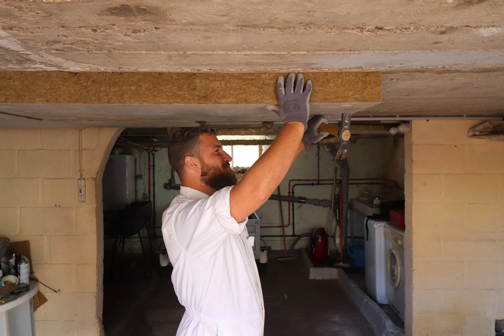 Artisan en combinaison blanche fixant un panneau de laine de roche sous la dalle béton d'une cave — isolation plafond des caves