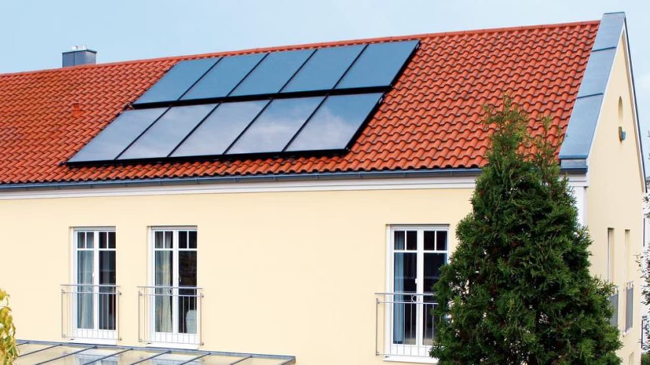 Dix panneaux solaires noirs intégrés en deux rangées dans une toiture en tuiles rouges d'une maison à façade jaune, avec un conifère au premier plan