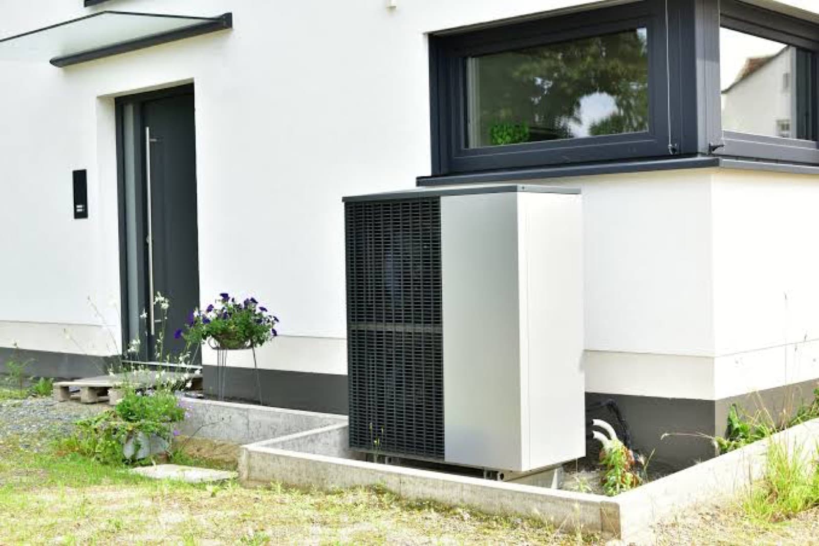 Unité extérieure blanche d'une pompe à chaleur air-eau installée contre la façade d'une maison, entourée de plantes