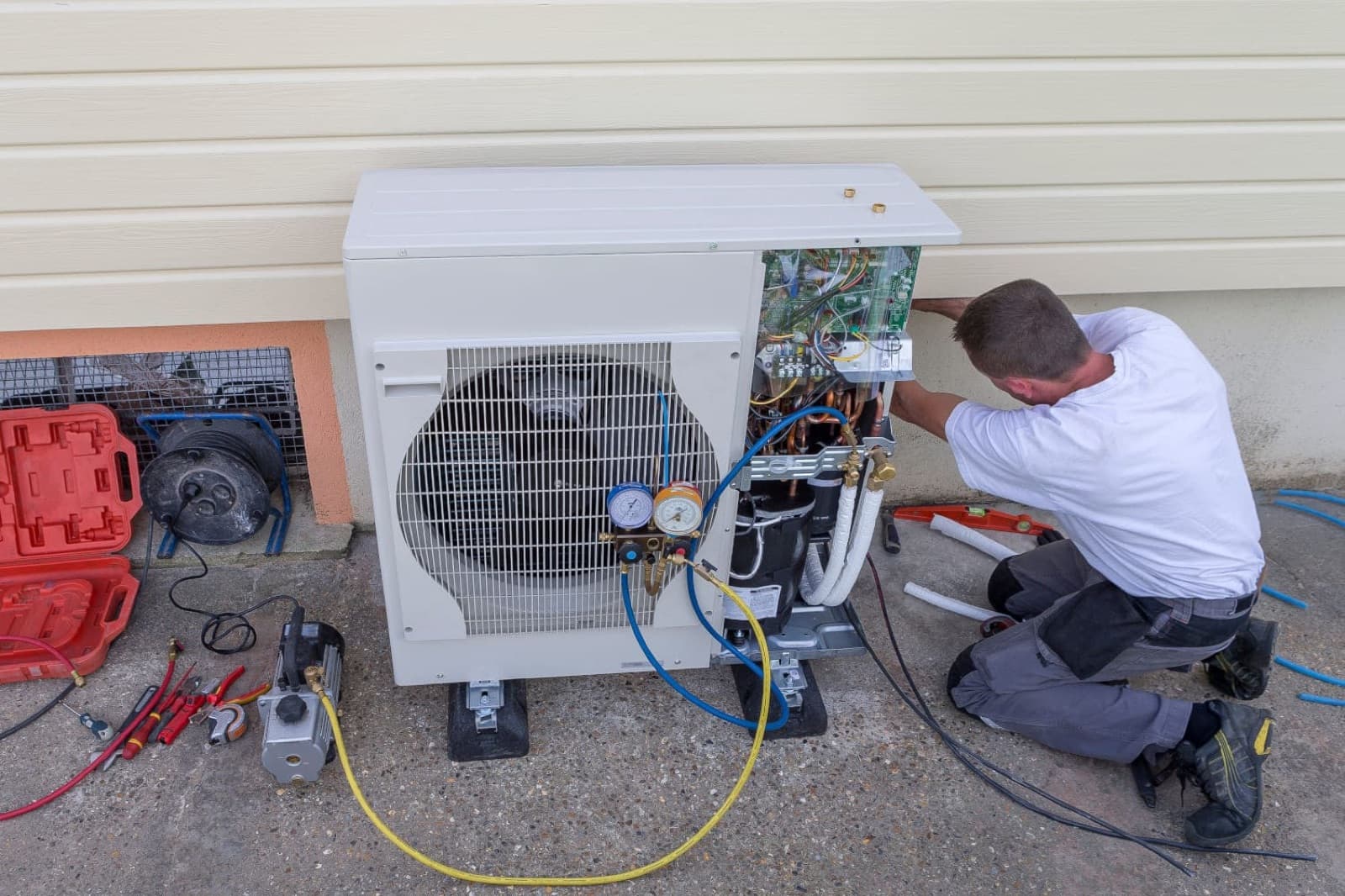 Technicien contrôlant les manomètres du circuit frigorifique d'une pompe à chaleur air-eau, boîtier électronique ouvert, outils de vide et raccords visibles