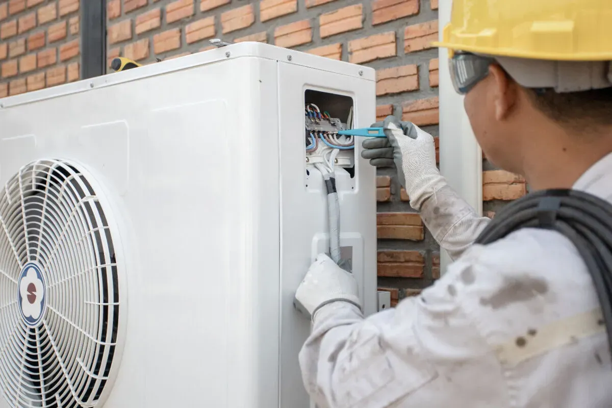 Technicien en casque de chantier contrôlant le tableau électrique d'une unité extérieure de pompe à chaleur lors d'un entretien annuel