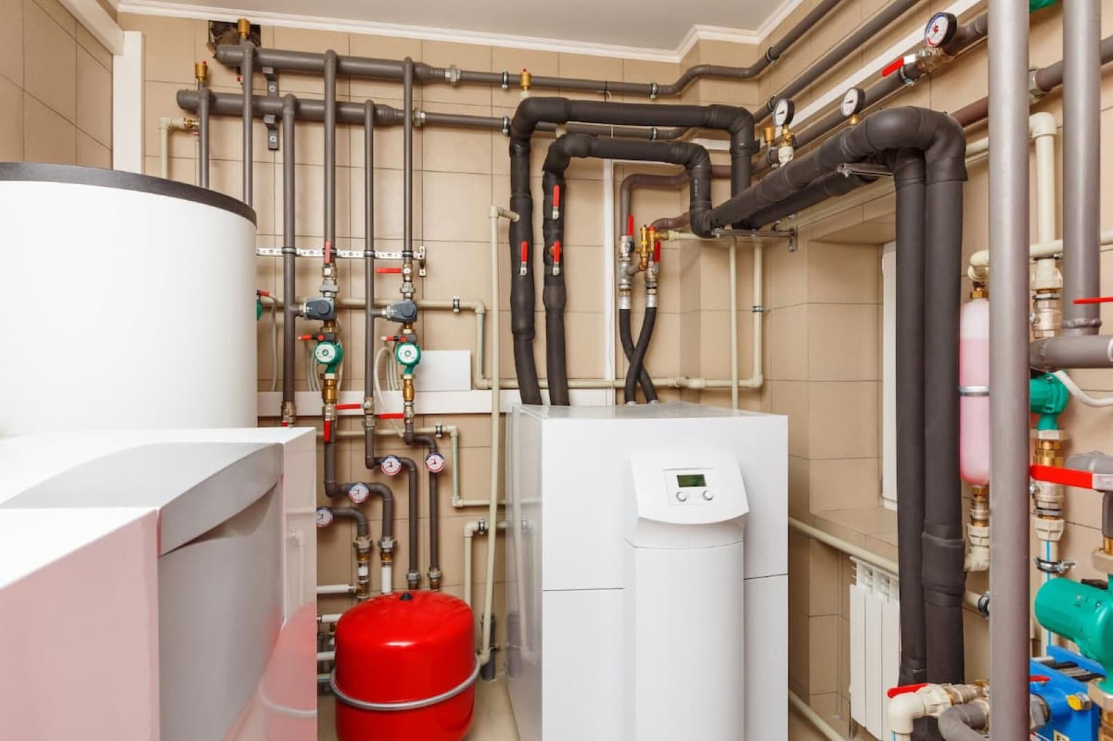 Unité intérieure blanche d'une pompe à chaleur géothermique installée dans un local technique, entourée de tuyaux, vase d'expansion rouge et manomètres