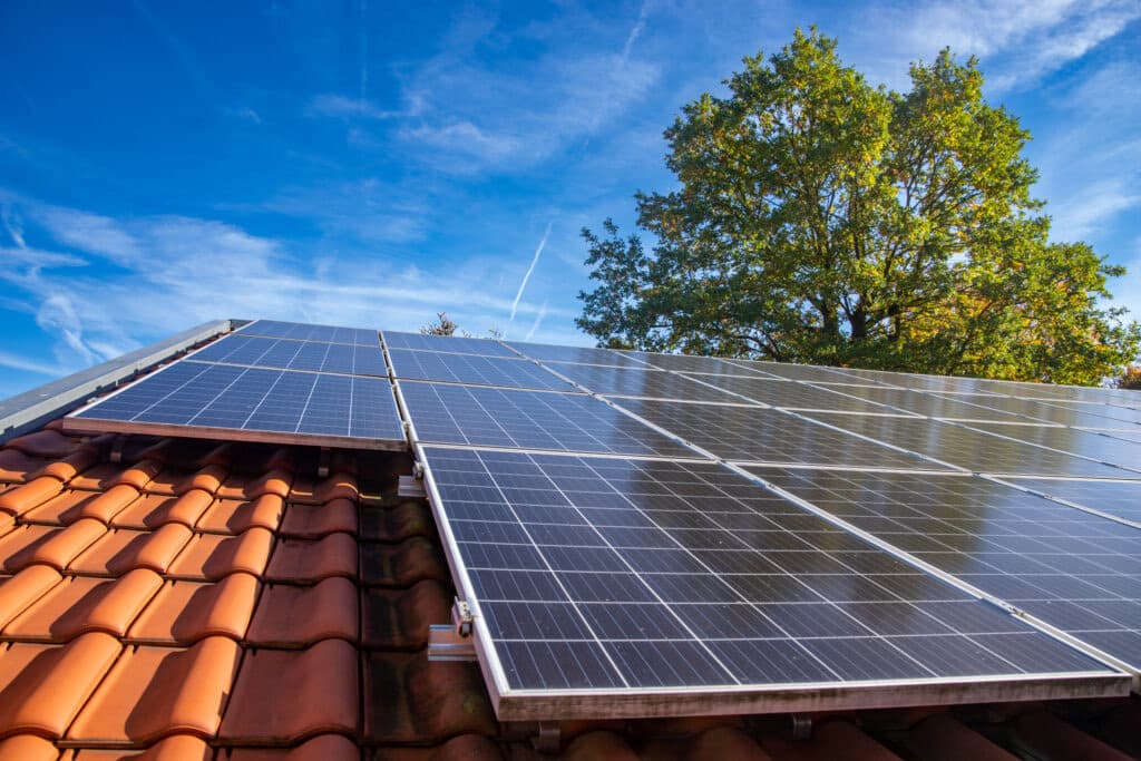 Rangées de panneaux photovoltaïques fixés sur un toit en tuiles mécaniques rouges, sous un ciel bleu, avec un grand chêne en arrière-plan