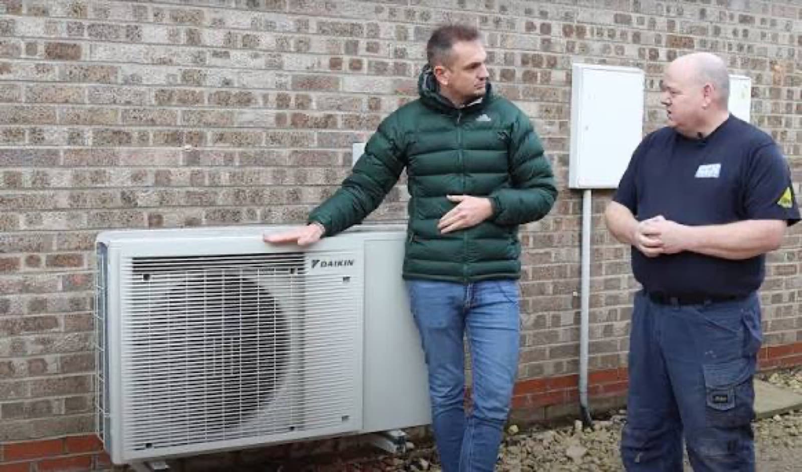 Un artisan chauffagiste explique à un client le fonctionnement d'une pompe à chaleur devant l'unité extérieure installée contre un mur en briques