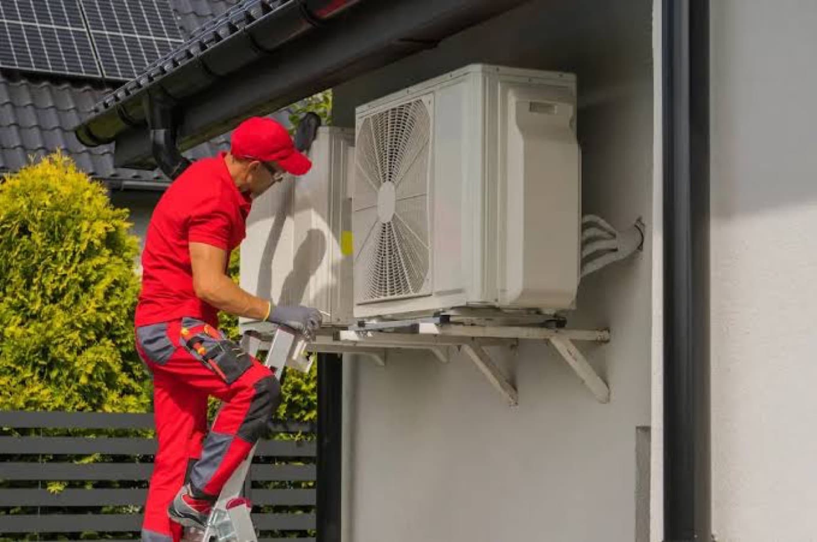 Technicien en tenue rouge installant sur une échelle l'unité extérieure d'une pompe à chaleur contre la façade d'une maison équipée de panneaux solaires