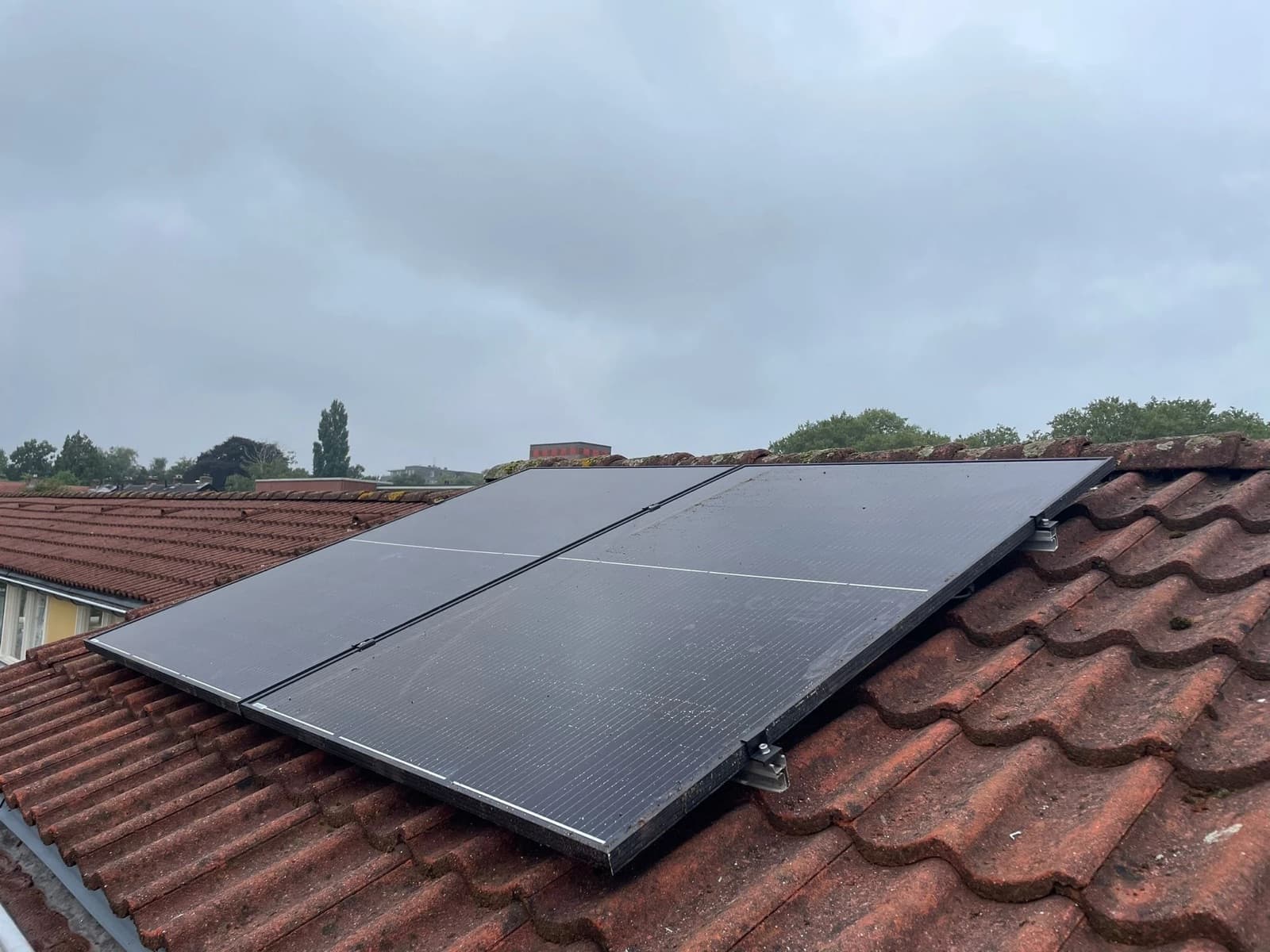 Deux panneaux solaires noirs fixés en surimposition sur une toiture en tuiles mécaniques rouges, avec les rails en aluminium et les crochets visibles sur le côté