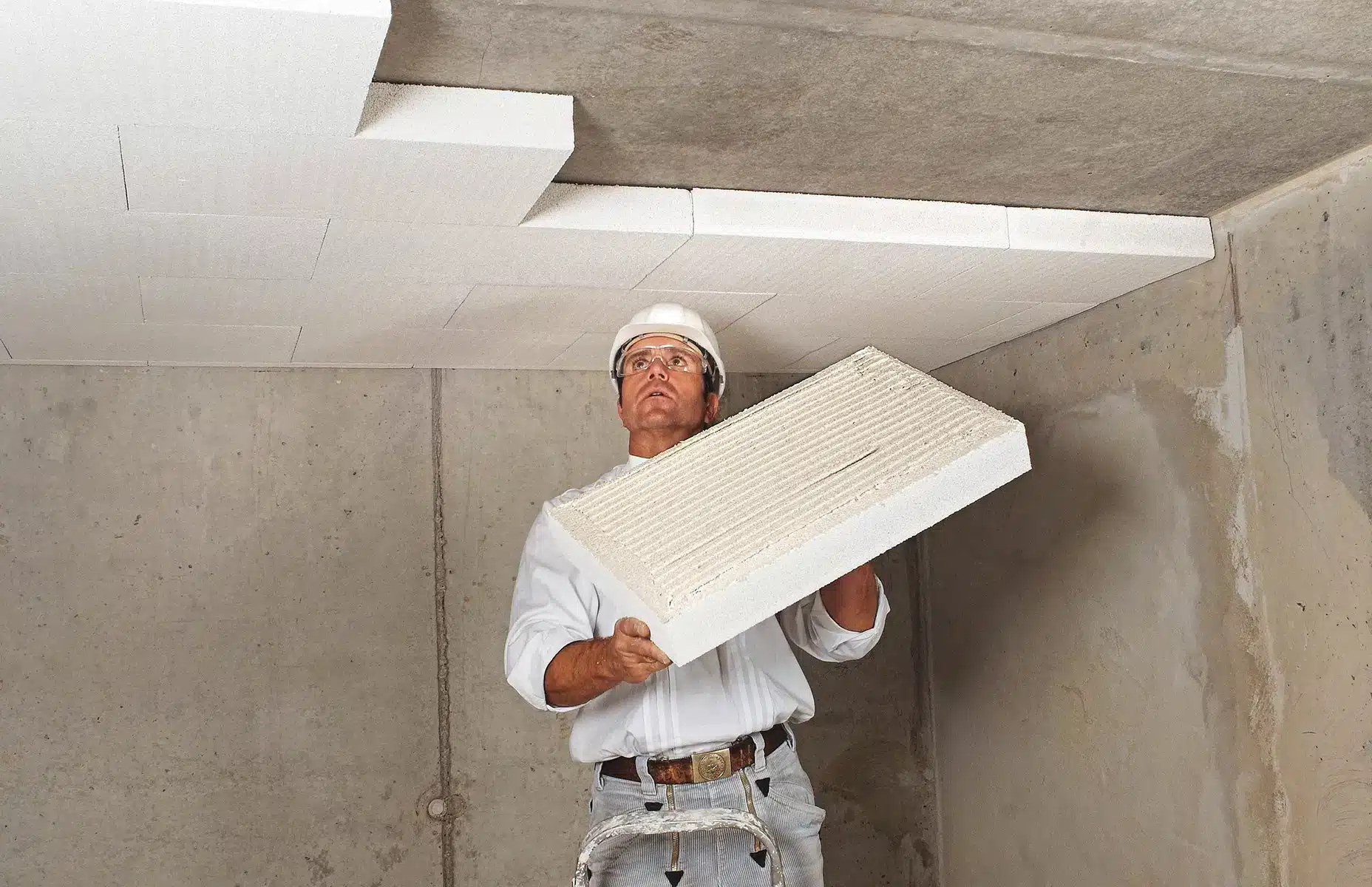 Artisan en casque de chantier posant des panneaux de polystyrène au plafond d'une cave en béton — isolation pour cave