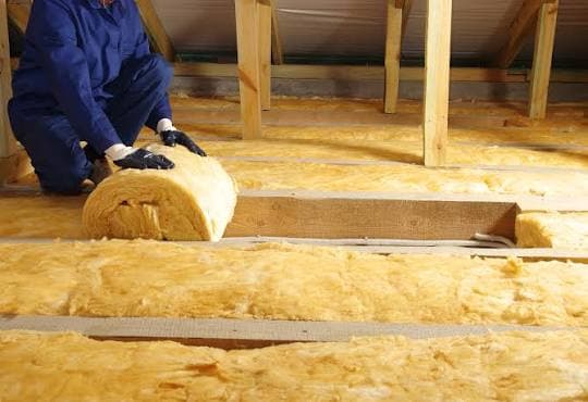 Pose manuelle d'un rouleau de laine de verre jaune entre les solives du plancher de combles perdus