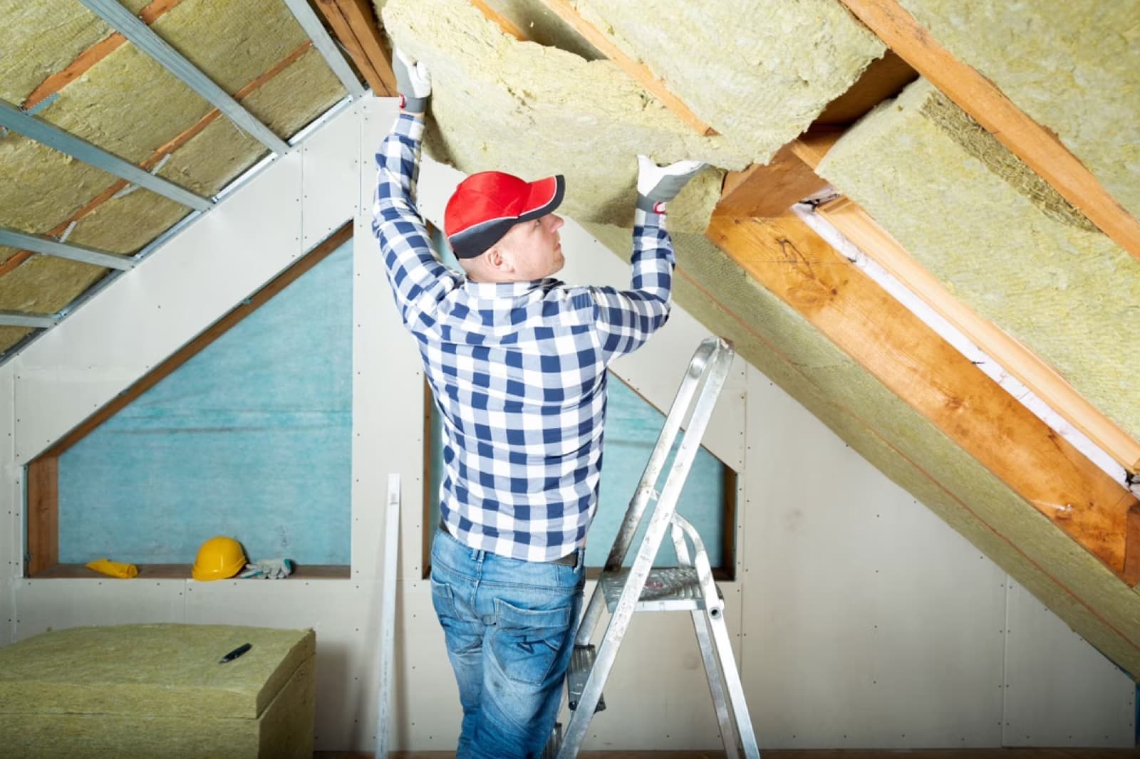 Artisan sur escabeau posant un panneau de laine de roche sous les rampants d'une toiture — chantier d'isolation des combles