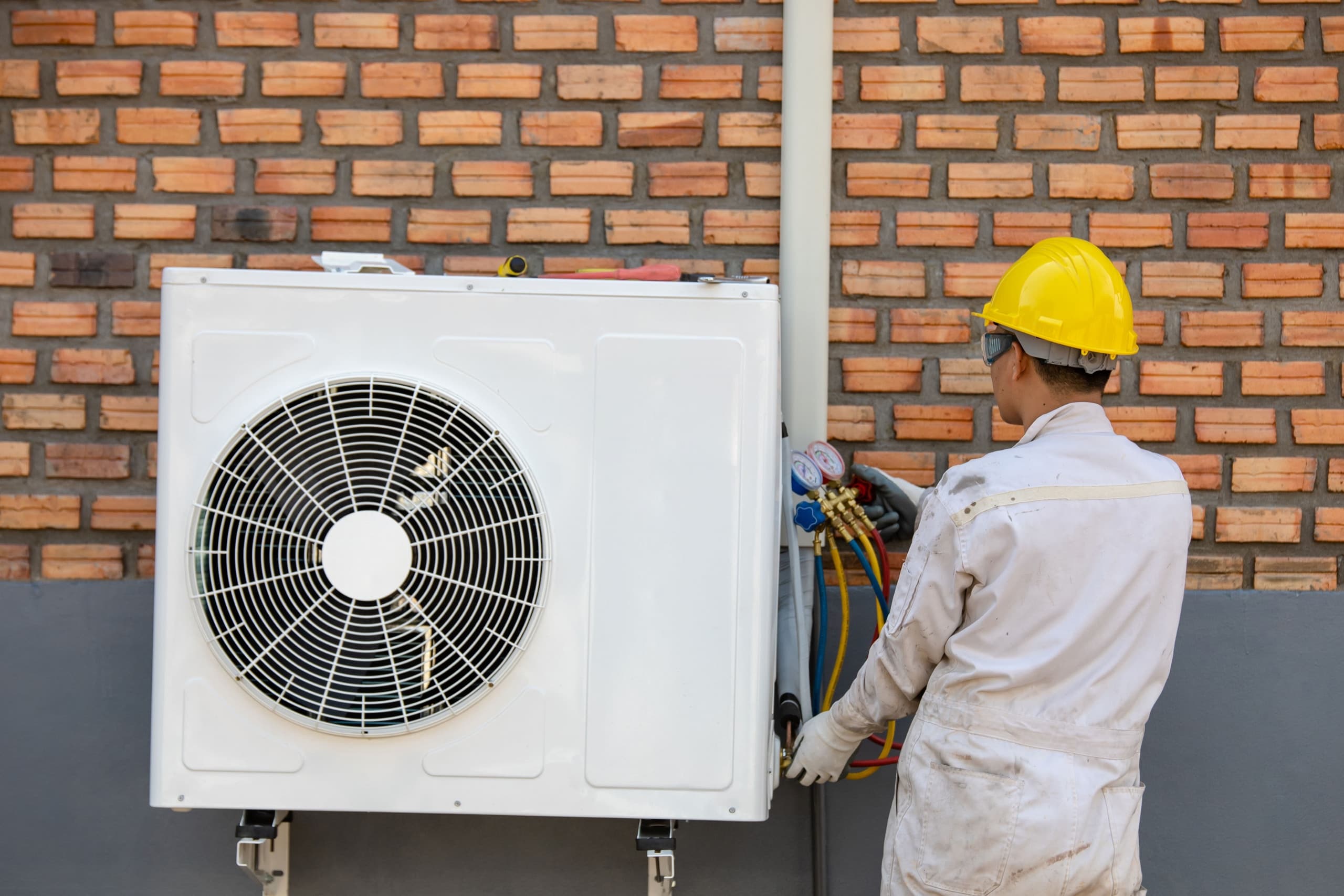 Technicien RGE en combinaison blanche et casque jaune raccordant une pompe à chaleur murale — installation professionnelle