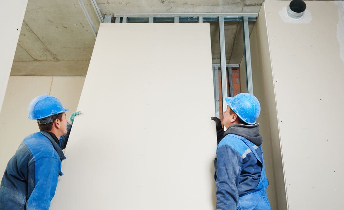 Deux artisans posant une plaque de plâtre isolante de grande dimension sur une ossature métallique au plafond, pour l'isolation thermique d'un logement en rénovation