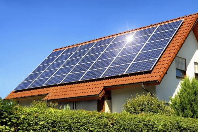 Maison individuelle dont tout le pan de toit en tuiles rouges est couvert de panneaux solaires bleus, au-dessus d'une haie, sous un ciel bleu