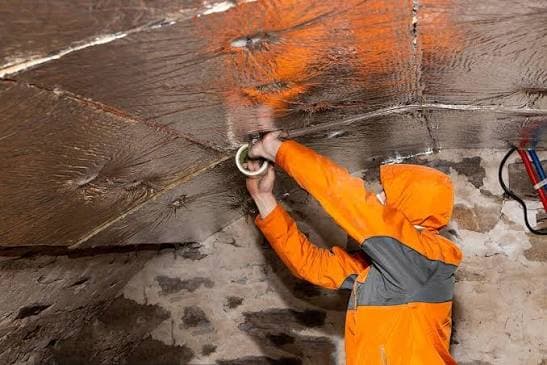 Technicien en combinaison orange appliquant une membrane d'étanchéité sur un mur de cave en pierre pour traiter les problèmes d'humidité avant l'isolation